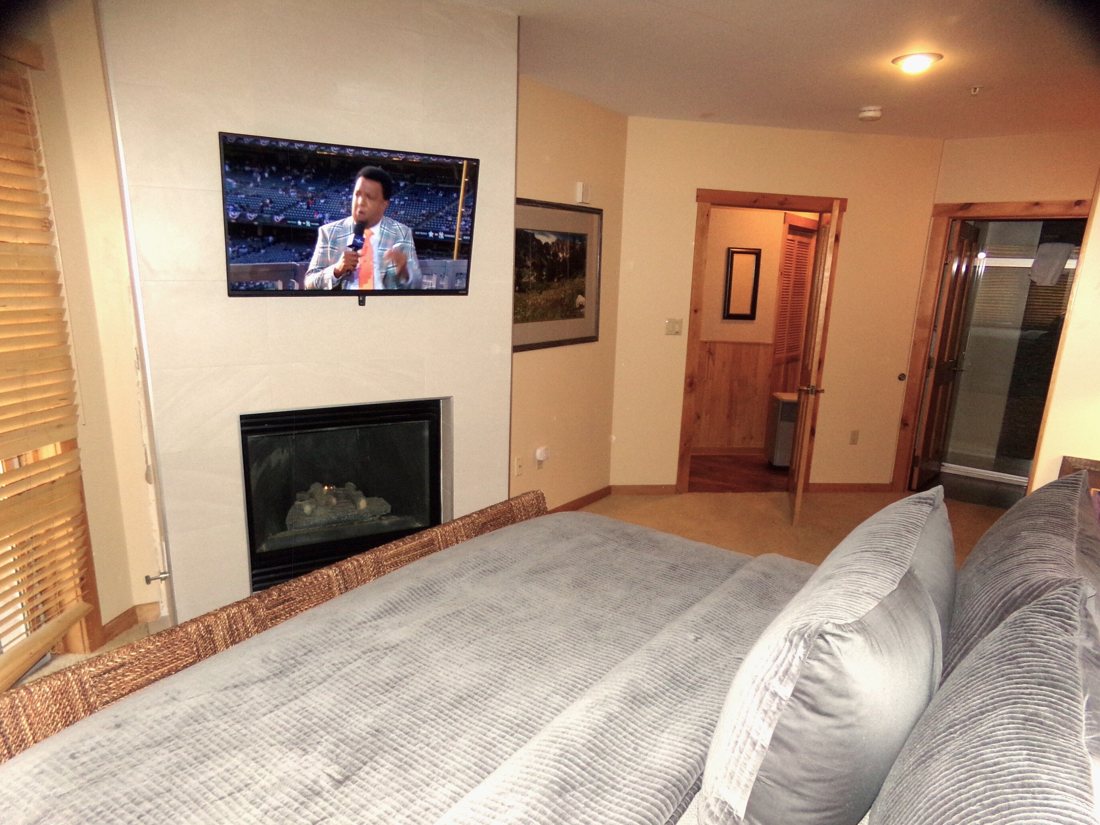 Un dormitorio con televisión montada en la pared, chimenea, cama con almohadas y puerta que da a otra habitación.