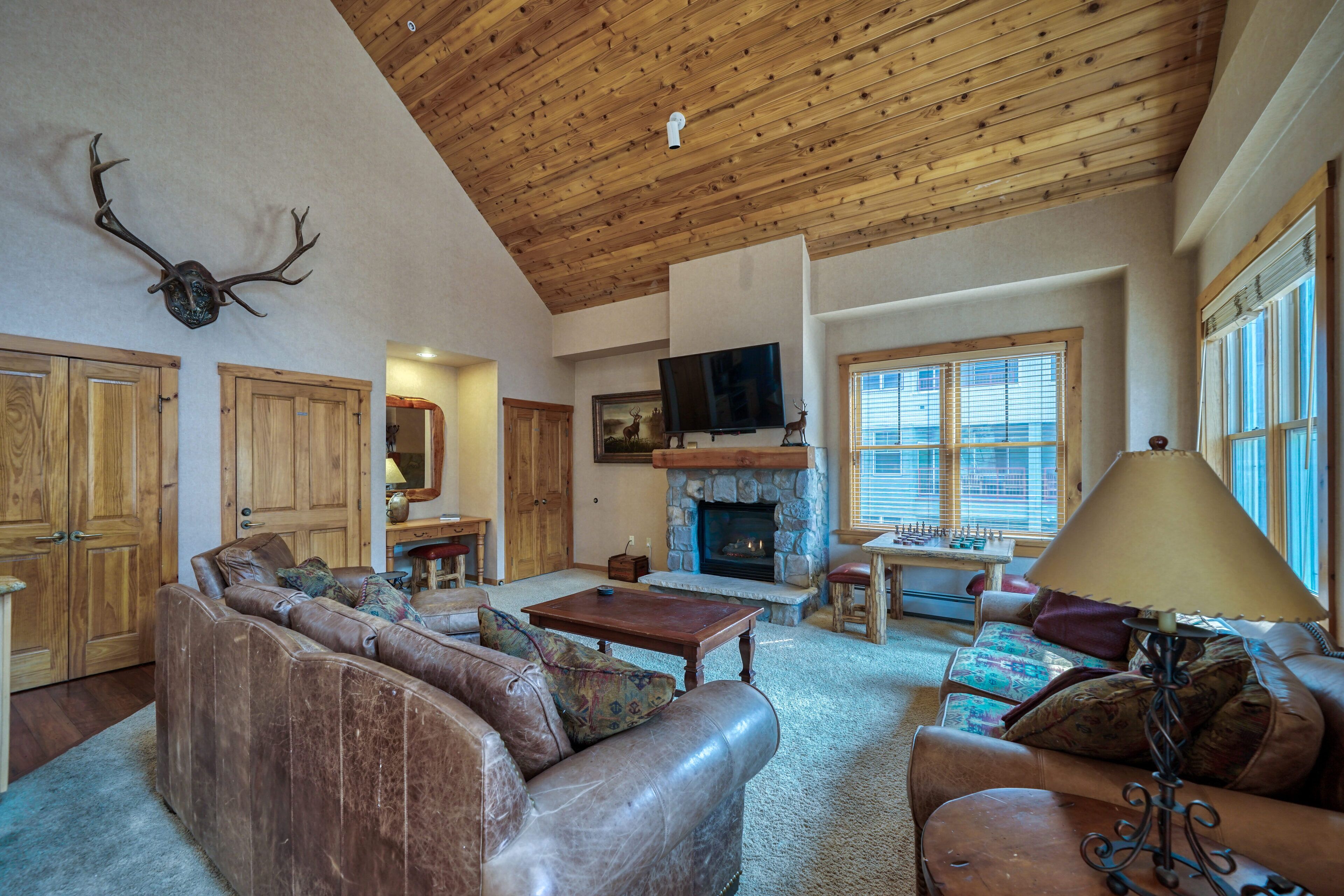 A spacious living room with a high wooden ceiling, a stone fireplace, leather sofas, and a television mounted on the wall.