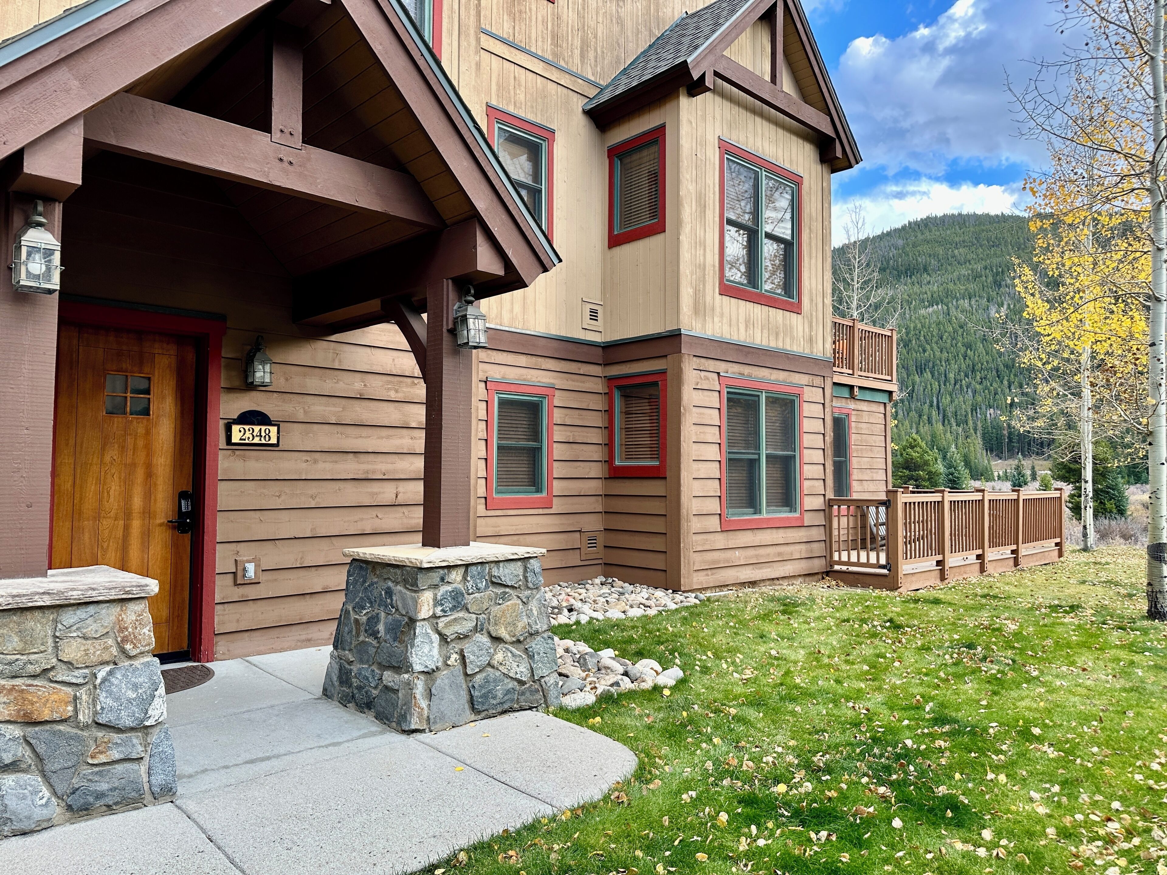 A two-story wooden house with a stone foundation, a number '2348' on the door, and a view of a forested mountain range.