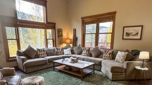 A cozy living room with a sectional sofa, a coffee table, and a rug. There are large windows with a view of trees and mountains. - Private Garage/Walk to the Gondola in 2 Minutes/Pool and Hot Tub/Fitness Room (Keystone)