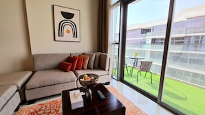 Ein modernes Wohnzimmer mit einem grauen Sofa, roten Kissen, einem Couchtisch mit Schale und Fernbedienung und einem großen Fenster mit Blick auf eine grüne Außenanlage.