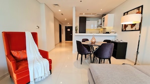 Ein modernes Wohnzimmer mit einem roten Sessel, einem Esstisch mit vier Stühlen und einer Couch. Im Hintergrund ist ein Küchenbereich mit weißen Schränken und einer Pendelleuchte zu sehen.