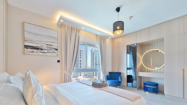A modern hotel room with a large bed, a blue chair, a round mirror, and a view of tall buildings. - Marco Polo - 1BR with Marina Views, Near Metro & Marina Mall (Dubai)