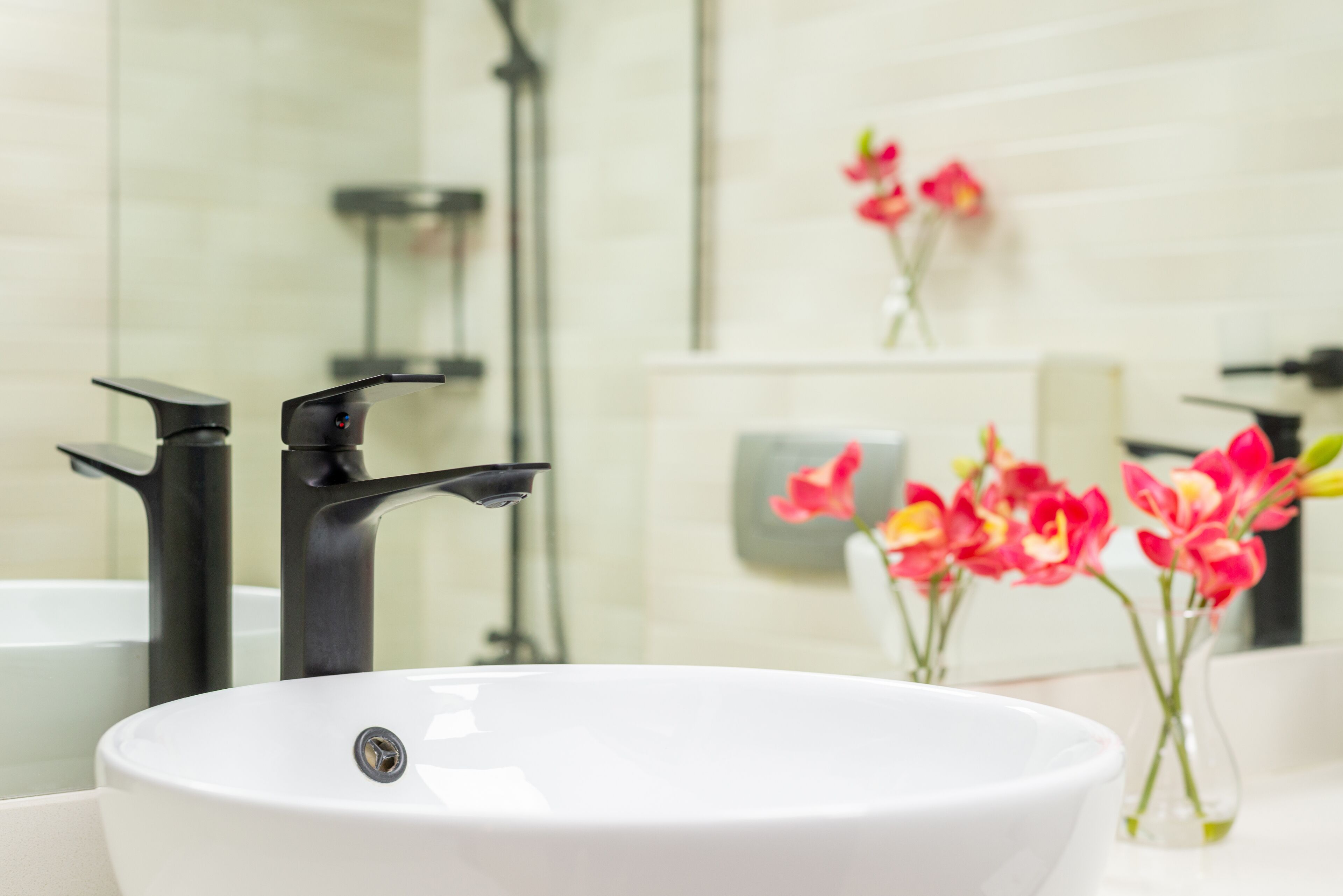 Ein weißes Waschbecken mit schwarzen Armaturen, eine Duschkabine aus Glas und rosa Blumen in einer Vase.