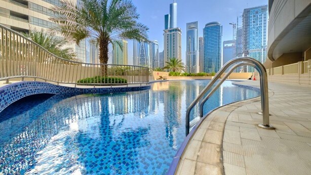 Una piscina con bordo piastrellato, circondata da una ringhiera metallica, palme e moderni grattacieli sullo sfondo.