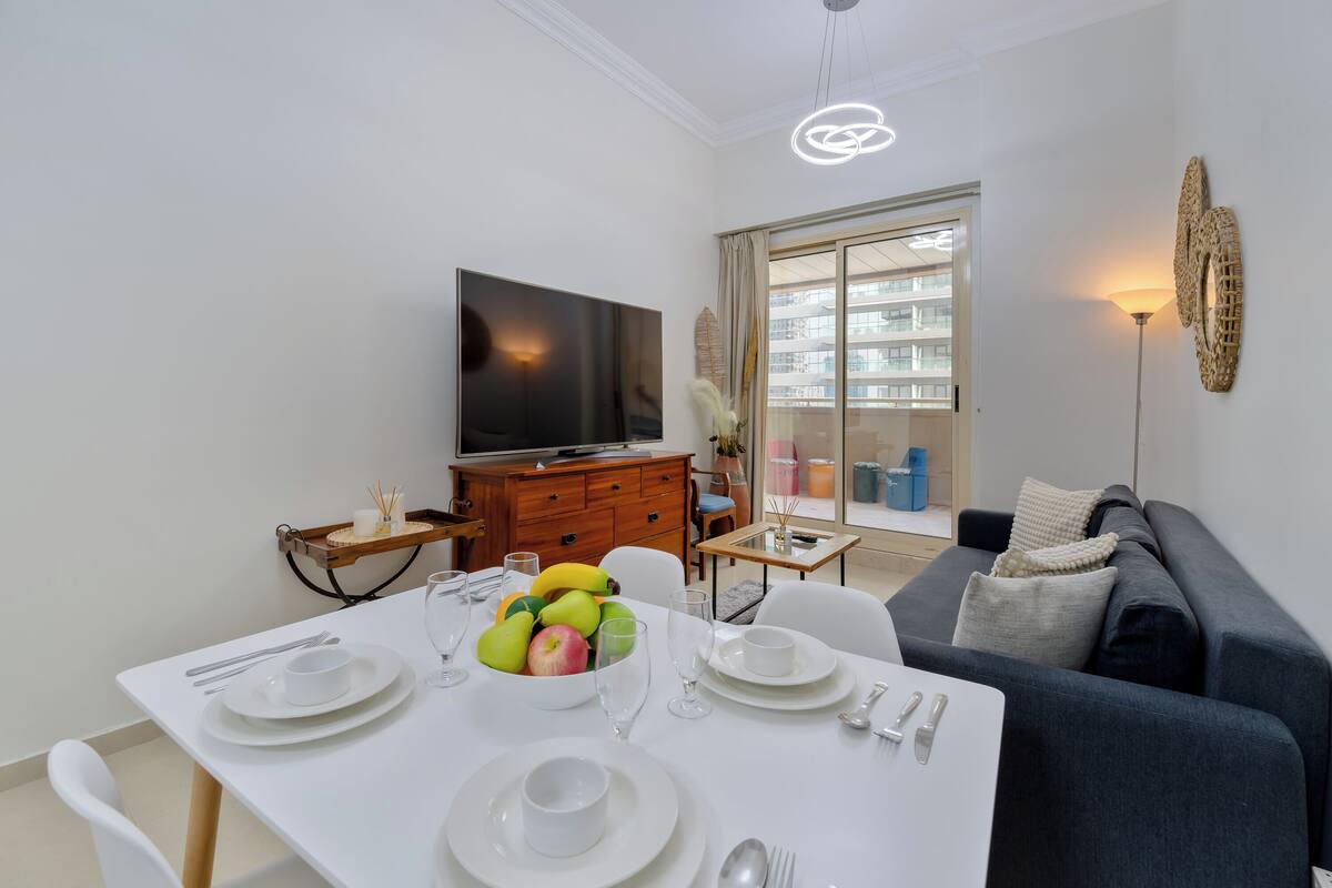 A dining area with a white table set for four, a television, and a sofa in the background.