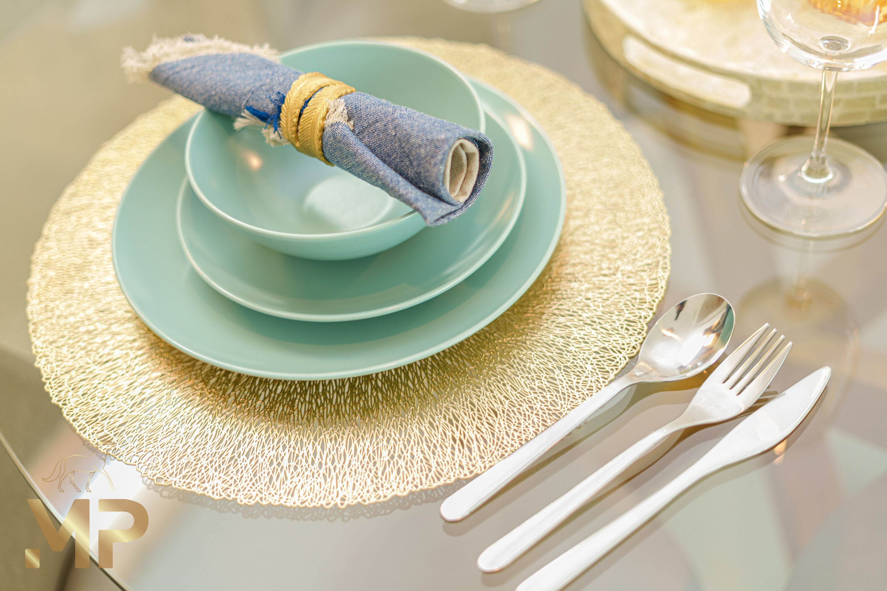 Une table dressée avec un bol bleu, une assiette décorative dorée et des couverts blancs.