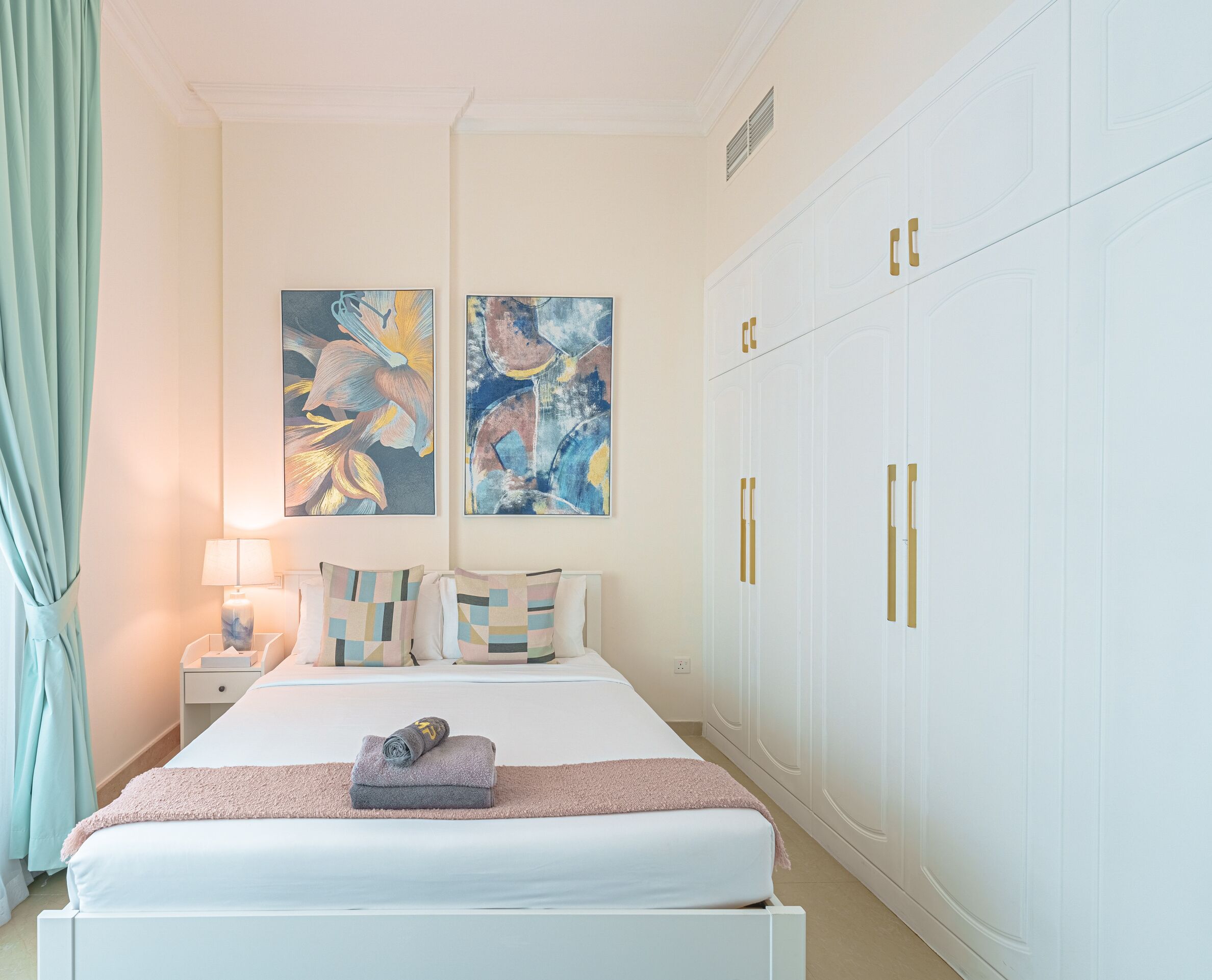 A bedroom with a bed, nightstands, a lamp, two abstract paintings, and a wardrobe with gold handles.