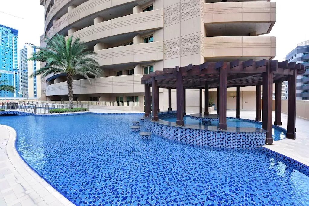 Une piscine aux carreaux de mosaïque bleus, entourée d’un bâtiment doté de nombreux balcons et d’un palmier.