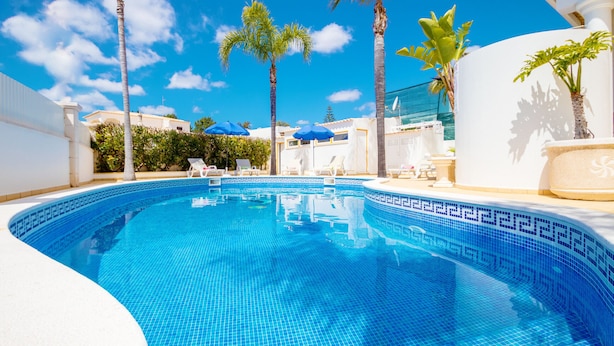 A swimming pool with blue mosaic tiles, surrounded by palm trees and white lounge chairs.