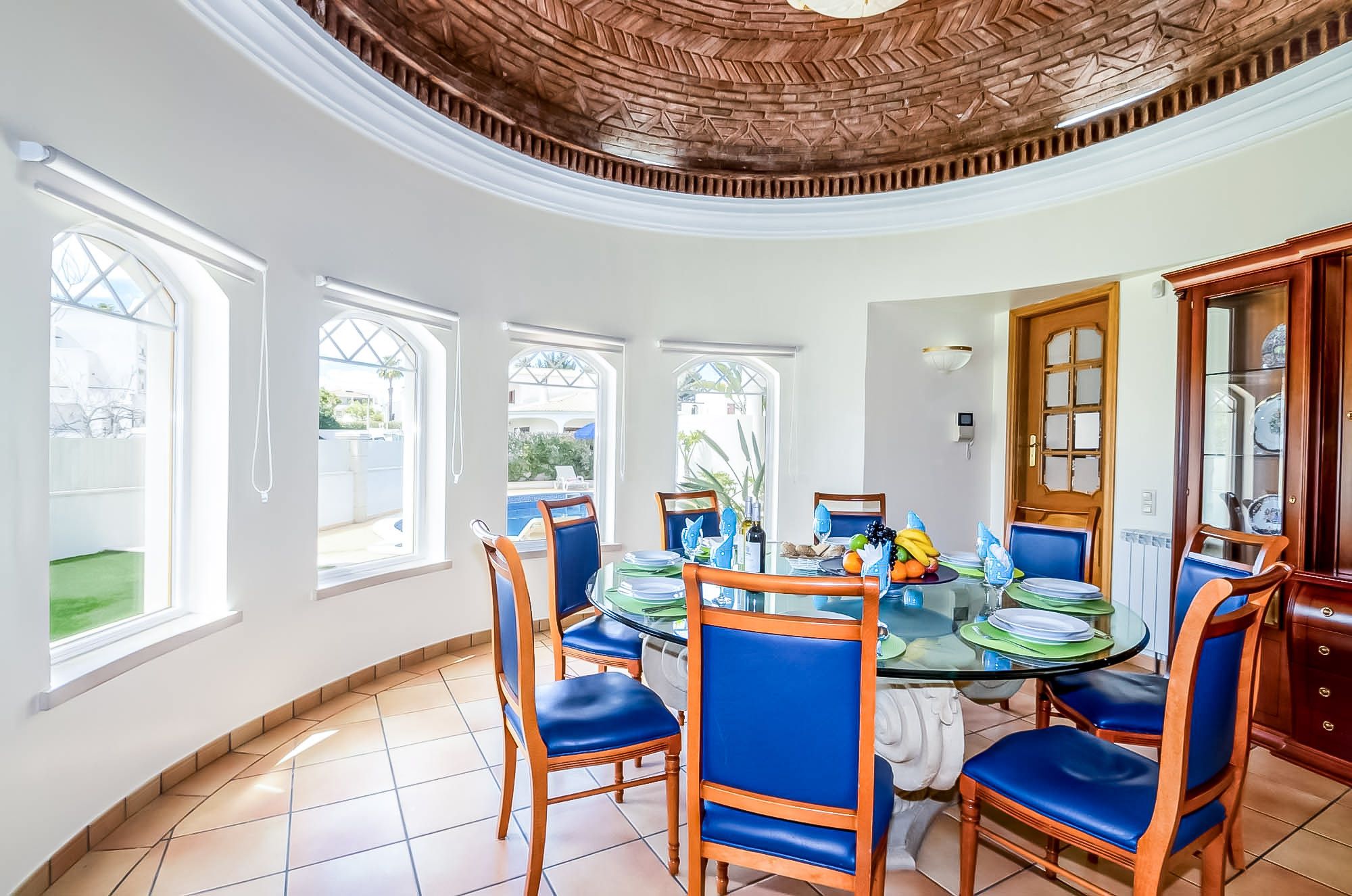 Un comedor con una mesa redonda de vidrio, sillas tapizadas en azul y vistas a una piscina por las ventanas.