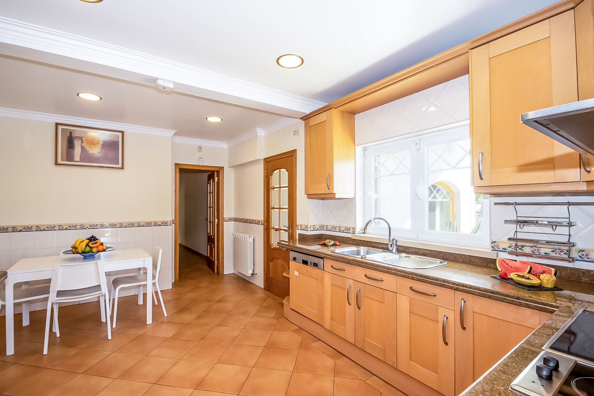 A kitchen with wooden cabinets, a dining table with chairs, a fruit bowl, and a window with a view.