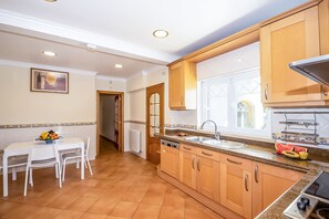 A kitchen with wooden cabinets, a dining table with chairs, a fruit bowl, and a window with a view. - Villa w/ heatable pool & BBQ, Albufeira (Albufeira)
