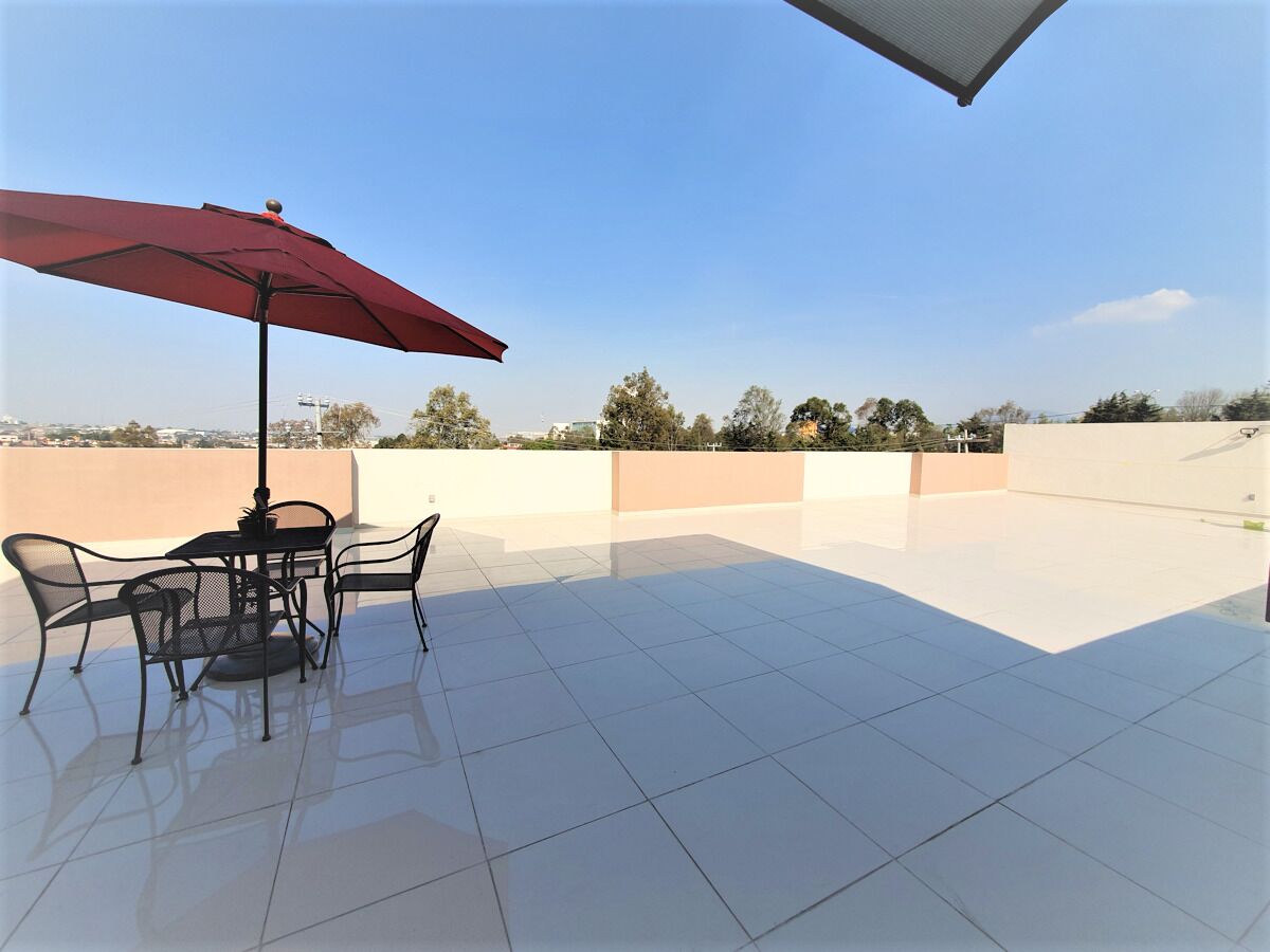 A rooftop terrace with a table and chairs under a red umbrella, surrounded by a clear sky and distant trees.