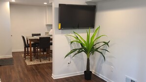 A modern living room with a flat-screen TV mounted on the wall, a dining area with a wooden table and chairs, and a potted plant. - Entire Lovely Apt* 5 Miles to EWR* Free Parking (Linden)