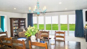 A dining area with a wooden table, chairs, a chandelier, a bookshelf, and large windows with a view of a green field. - Ocean Front, Amazing Views! Hot Tub! Dog Friendly! (Yachats)