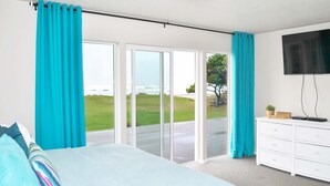 A bedroom with a large bed, a white dresser, a flat-screen TV, and a sliding glass door leading to a deck with a view of the ocean. - Ocean Front, Amazing Views! Hot Tub! Dog Friendly! (Yachats)