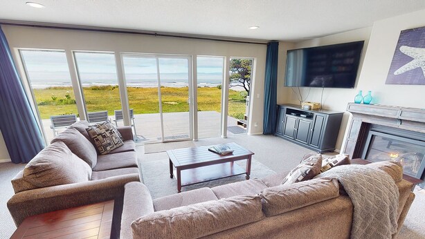 A living room with a sofa, coffee table, and a fireplace. There is a television mounted on the wall and a view of the outdoors through large windows. - Ocean Front, Amazing Views! Hot Tub! Dog Friendly! (Yachats)