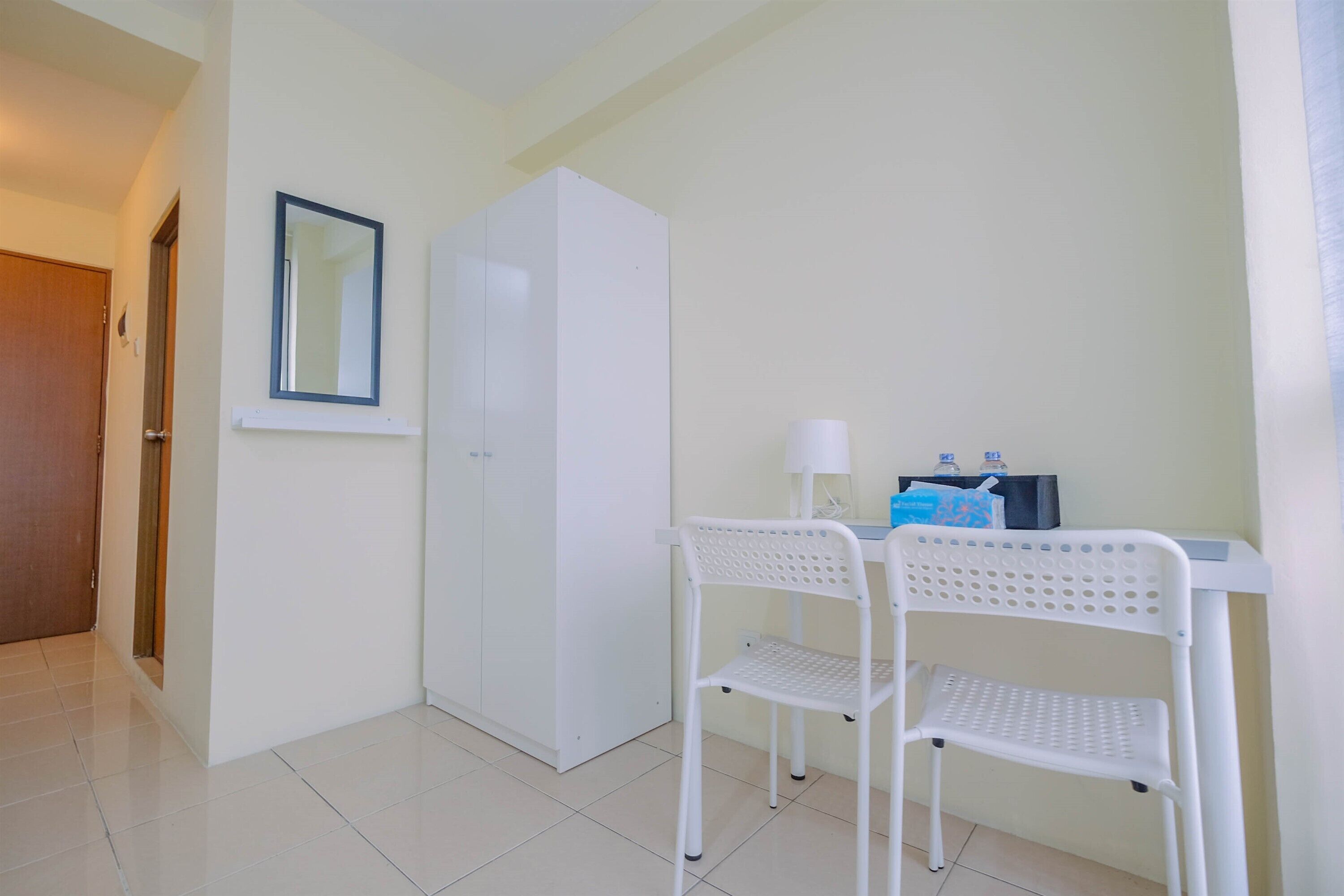 A small, clean room with a white wardrobe, a white chair, and a desk with a lamp.
