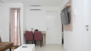 A compact living space with a sofa, a small dining table with two chairs, a TV mounted on the wall, and an air conditioning unit.