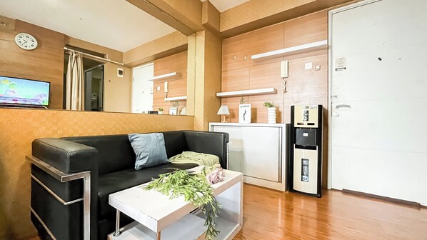 A modern living room with a black leather sofa, a white coffee table with a bouquet of flowers, a small white cabinet, a wall-mounted clock, a television, a refrigerator, and a door with a handle. - Compact and Comfy 2BR Bassura City Apartment (Jakarta)