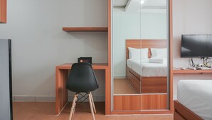 A hotel room with a wooden desk, a black chair, a bed with white bedding, a television, and a sliding door closet. - Best Homey Elegant Studio Room at Amethyst Apartment (Jakarta)