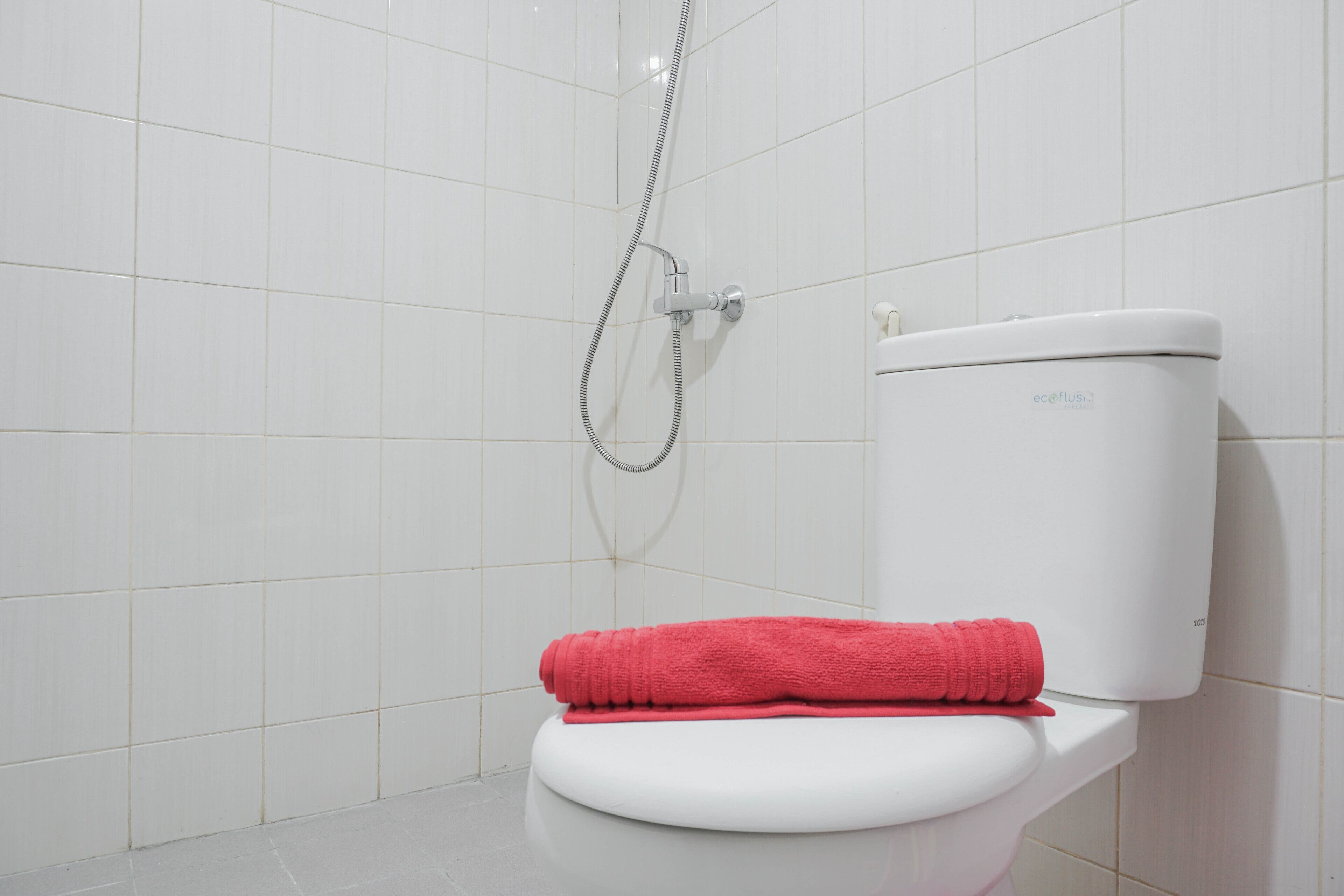 A white toilet with a red towel on top, a showerhead, and a white tiled wall.