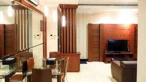A modern living room with a glass dining table, leather chairs, a wooden entertainment unit with a flat-screen TV, and a built-in air conditioning unit. - 2BR Prestige Dharmawangsa Essence Apartment Suites (Jakarta)