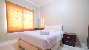 A neatly arranged bedroom with a large bed, a bedside table, a nightstand, and a window with blinds. - Strategic 2BR at Sudirman Park Apartment (Jakarta)