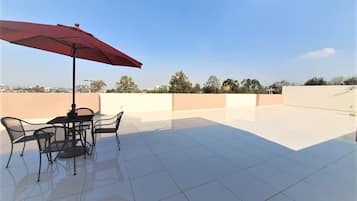 A rooftop terrace with a table and chairs under a red umbrella, surrounded by a clear sky and distant trees.