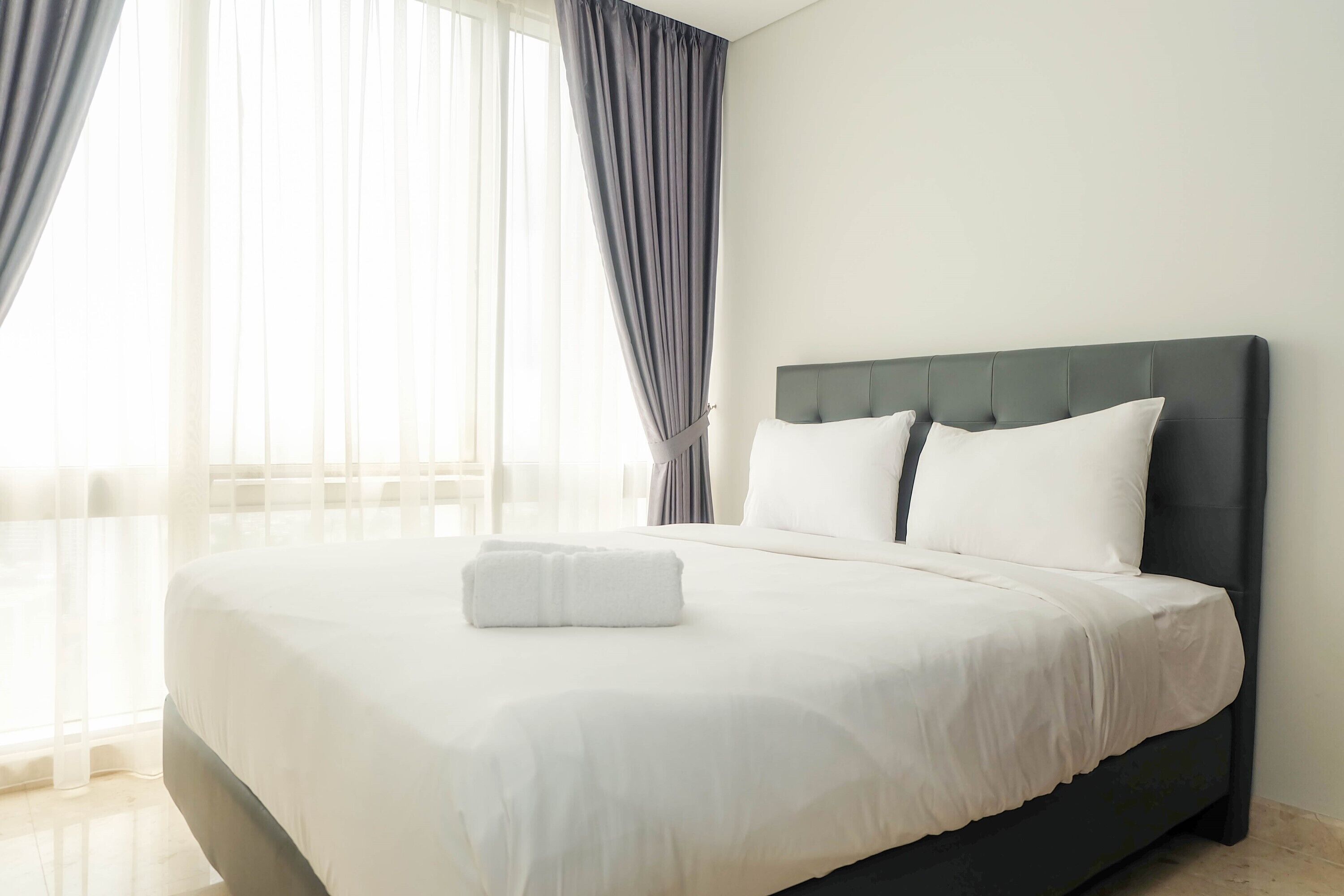 A neatly made bed with white linens and a folded towel, a dark headboard, and sheer curtains on a window.