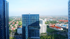 A high-rise building with a glass facade overlooking a cityscape with residential areas and a large building with a red roof. - Modern Studio Apartment at Tanglin Tower Supermall Mansion (Surabaya)