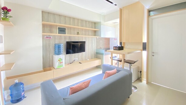 A modern living room with a grey sofa, a flat-screen TV mounted on the wall, a water dispenser, and a small table with a chair. - Cozy 2BR Apartment at Parahyangan Residence Bandung near UNPAR (Bandung)