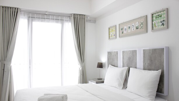 A hotel room with a large bed, a nightstand with a lamp, and a view of the outdoors through the window. - Modern Look Studio Apartment at Grand Asia Afrika (Bandung)