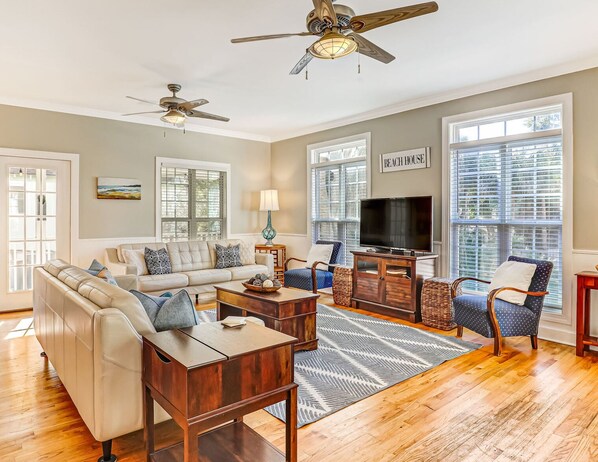 A spacious living room with a beige sofa, a wooden coffee table, and a TV. There are two ceiling fans, large windows with blinds, and a sign that reads "BEACH HOUSE".