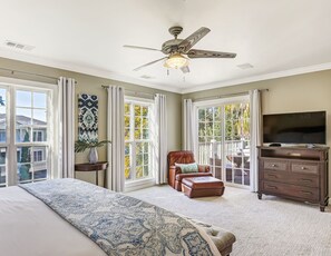 A bedroom with a large bed, a ceiling fan, a TV, a dresser, and a balcony with a view of trees.