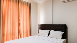 Una cama bien hecha con sábanas blancas y una toalla doblada, cabecera oscura y cortinas anaranjadas.
