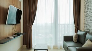 A modern living room with a flat-screen TV mounted on a wooden wall, a leather sofa, a small coffee table, and sheer curtains allowing natural light. - Comfort and Cozy 1BR at Tree Park City BSD Apartment (South Tangerang)