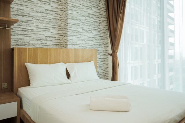 A hotel room with a bed, wooden headboard, white bedding, and a view of buildings through large windows. - Comfort and Cozy 1BR at Tree Park City BSD Apartment (South Tangerang)