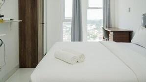 A hotel room with a large bed, a wooden wardrobe, a desk, and a view of the city through the window. - Cozy Studio at Amazana Serpong Apartment (South Tangerang)
