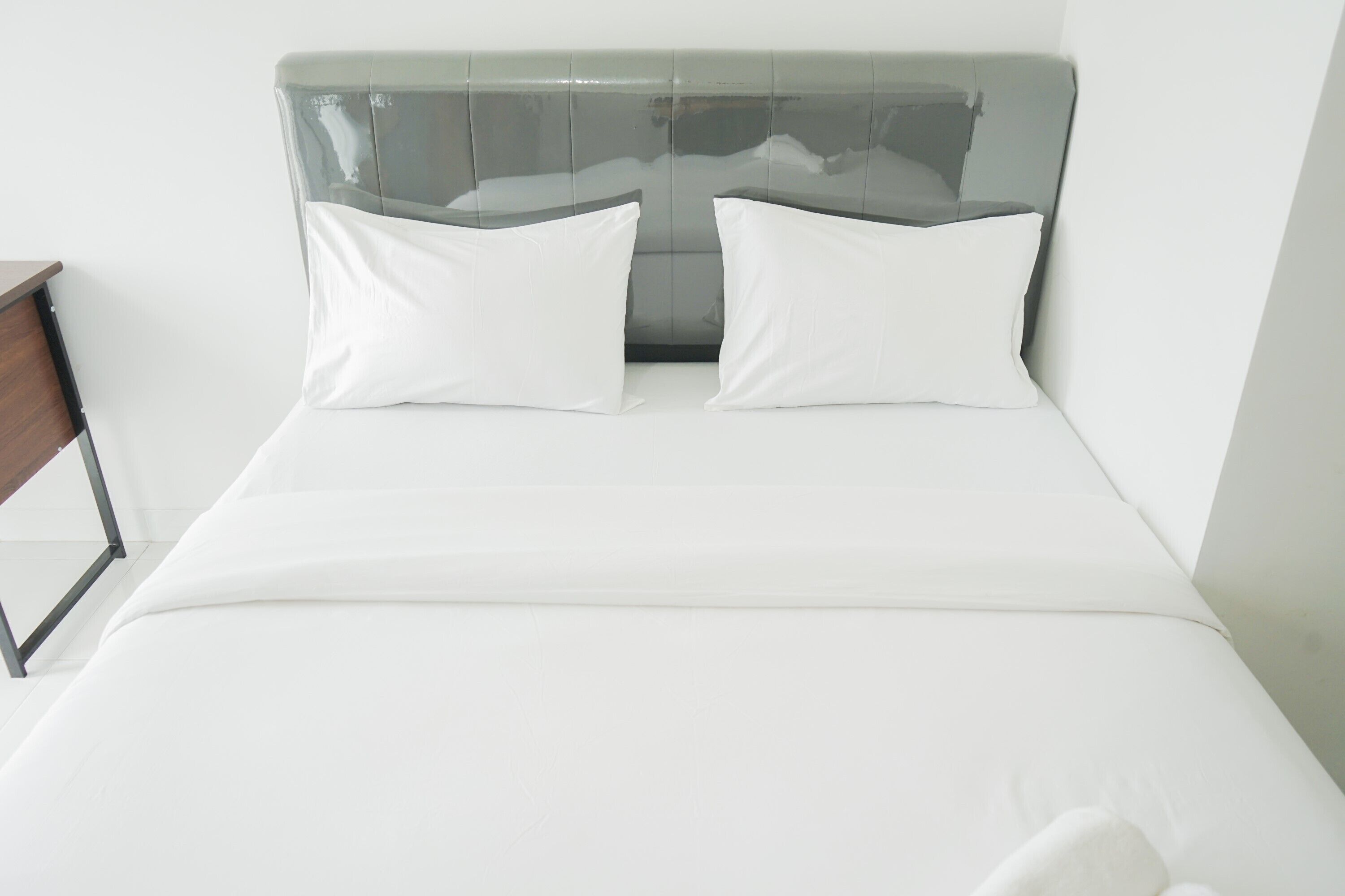 Una cama con cabecera de vidrio, dos almohadas blancas y una mesita de noche de madera a la izquierda.