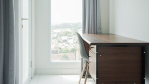 A room with a wooden desk, a chair, a window with curtains, and a view of buildings outside. - Cozy Studio at Amazana Serpong Apartment (South Tangerang)