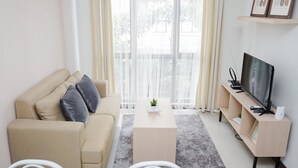A modern living room with a sofa, a coffee table, a TV on a wooden stand, and a large window with sheer curtains. - Comfortable and Best Choice 1BR at Asatti Apartment (Pagedangan)