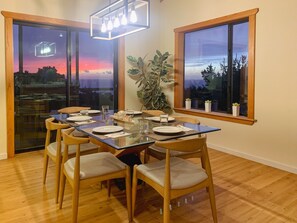 A dining area with a glass table, wooden chairs, and a view of a sunset through a glass door.