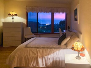 A bedroom with a bed, a chair, a desk, and a window with a view of the sea.