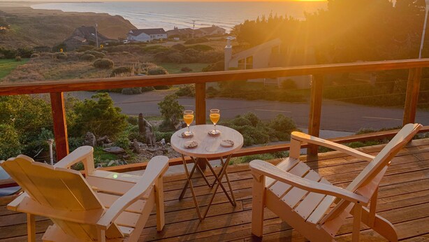 Een houten terras met twee Adirondack-stoelen en een kleine ronde tafel met drankjes, met uitzicht op een kustlandschap met huizen en de oceaan tijdens zonsondergang.