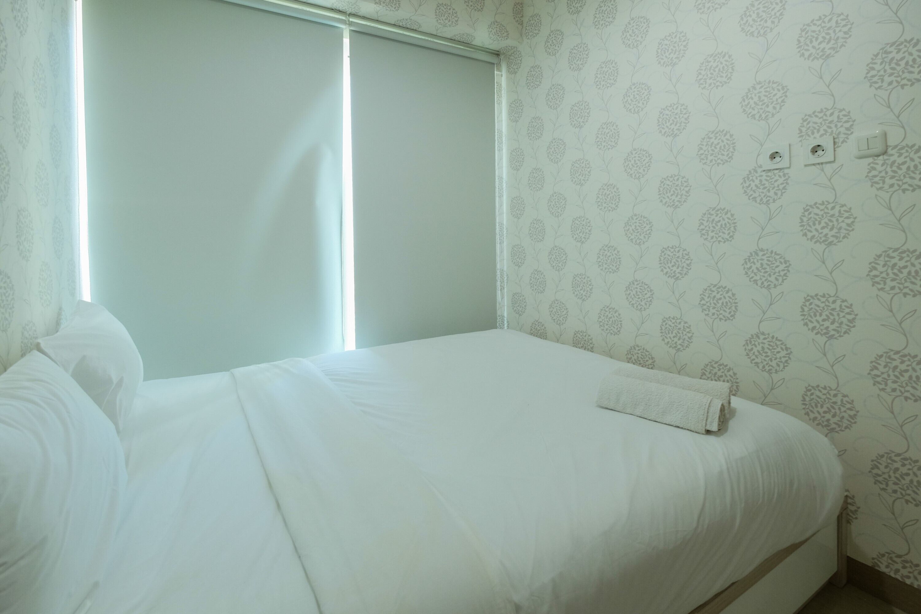 A neatly made bed with white bedding, a window with blinds, and a wall with a floral pattern.