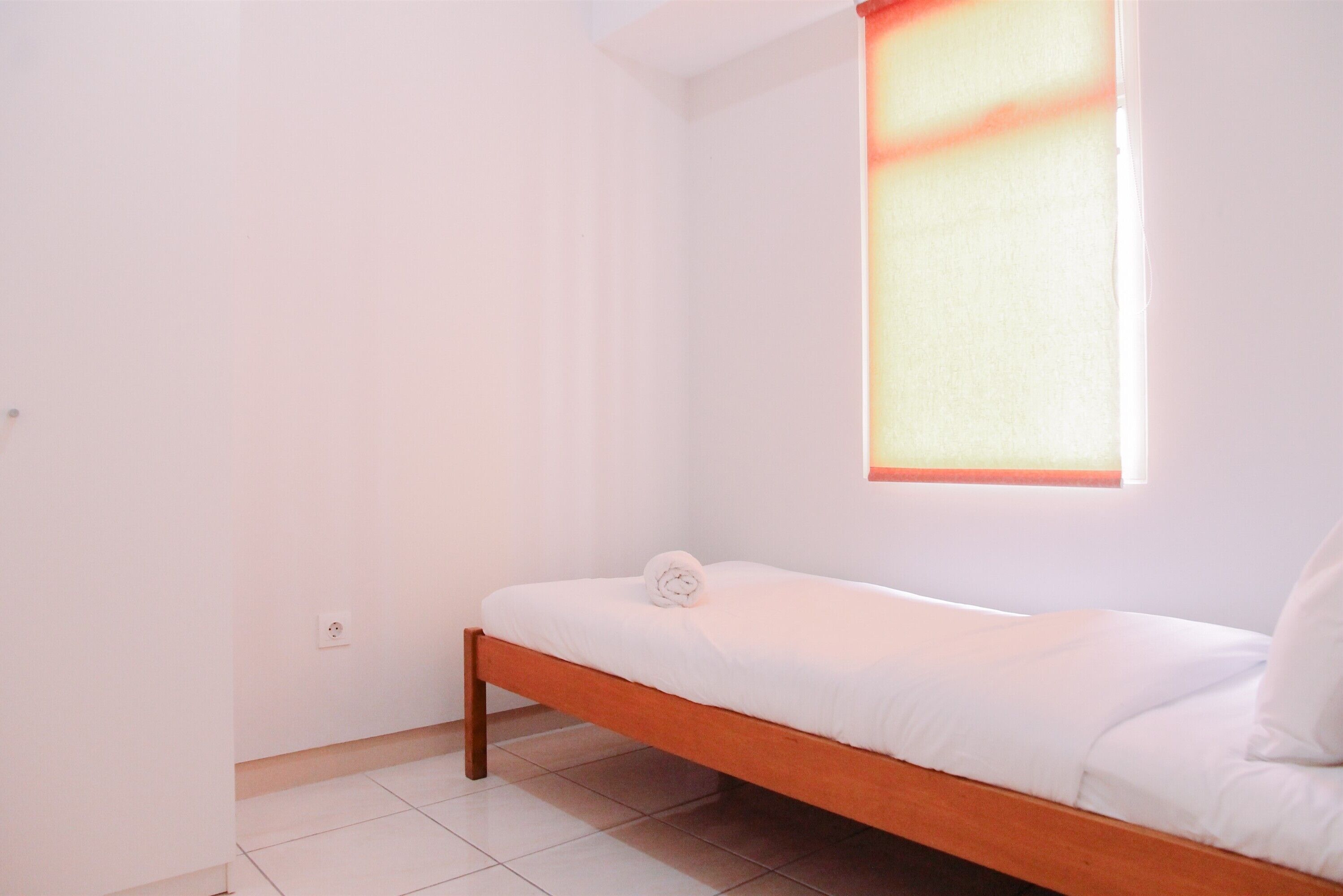 Une petite chambre avec un lit simple, un cadre en bois, du linge de lit blanc et une fenêtre avec un store de couleur claire.