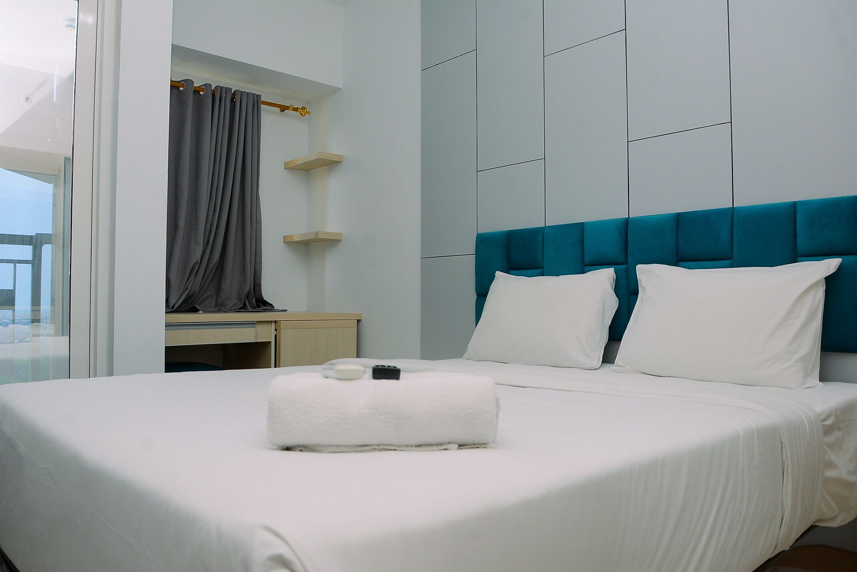 A hotel room with a large bed, a blue headboard, a white bedside table, and a grey curtain.