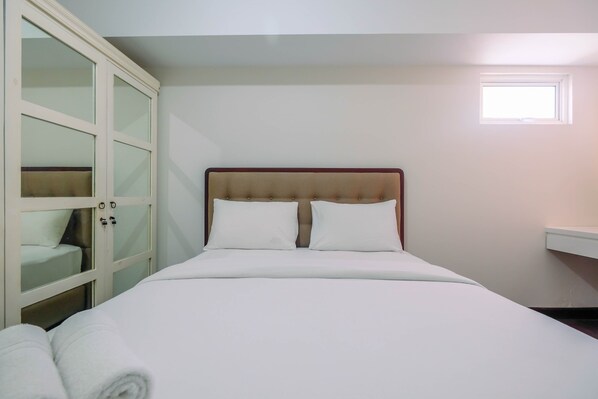 A neatly made bed with white linens, a wooden headboard, and a window with blinds.